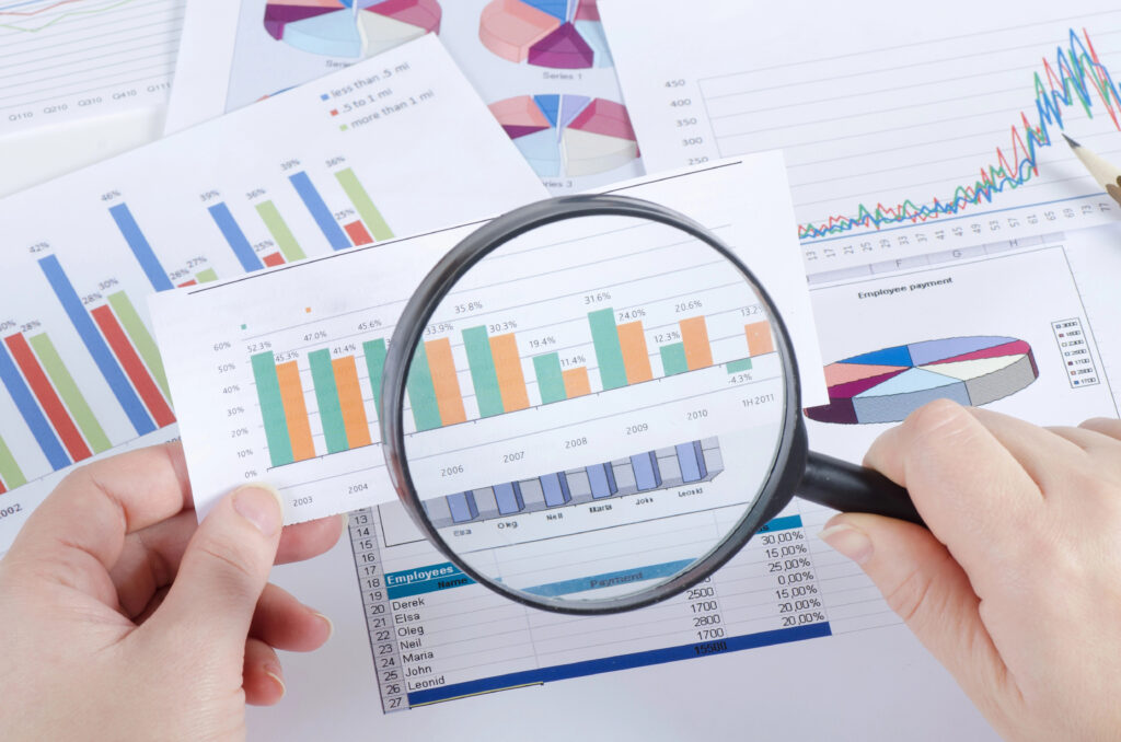 A person's hand holding a magnifying glass over printed bar and pie charts, focusing on analyzing data.